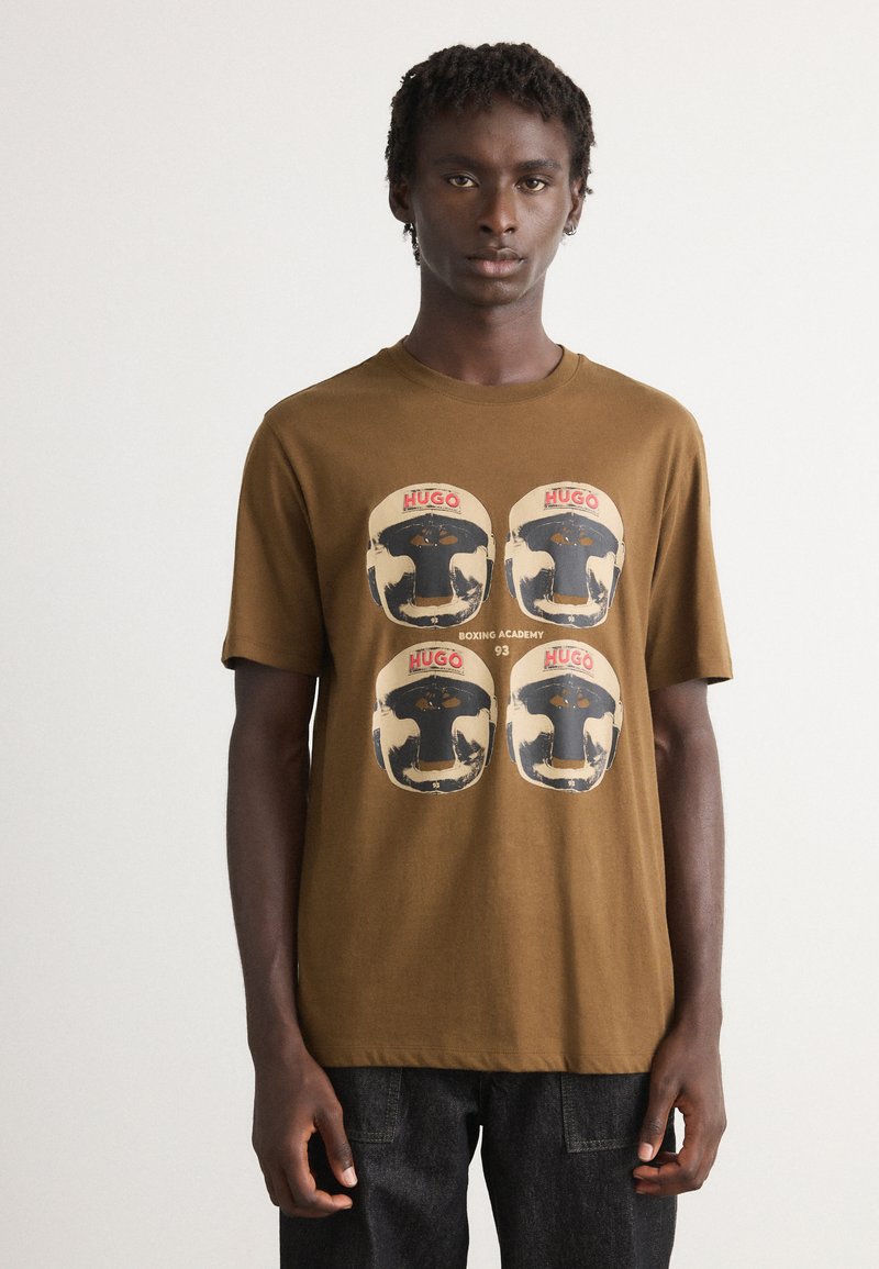 Junger Mann in einem braunen T-Shirt mit vier Boxhelm-Grafiken und dem Text "HUGO Boxing Academy 93", kombiniert mit dunkler Jeans, der nach vorne blickt.