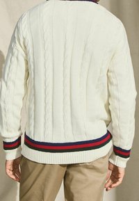 Mann in einem weißen Zopfmusterpullover mit rot, grün und marineblauen gestreiften Säumen und Bündchen, der vor einem beigen Hintergrund steht.