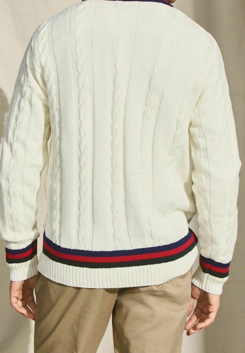 Mann in einem weißen Zopfmusterpullover mit rot, grün und marineblauen gestreiften Säumen und Bündchen, der vor einem beigen Hintergrund steht.