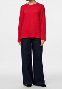 Czerwony sweter z dzianiny z okrągłym dekoltem, opadającymi ramionami i ściągaczami, zestawiony z ciemnoniebieskimi spodniami o szerokich nogawkach. Prosty design i faktura.
