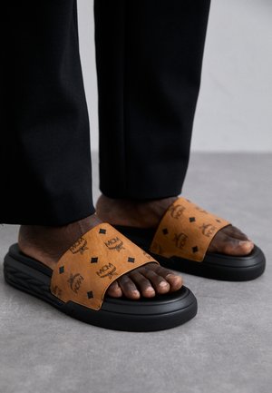 Brown slide sandals with black soles, featuring a patterned strap showcasing logos and diamond shapes. Textured surface, relaxed fit.