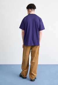 Purple cotton T-shirt with a relaxed fit, paired with light brown corduroy pants. The background features a blue carpet and neutral wall.