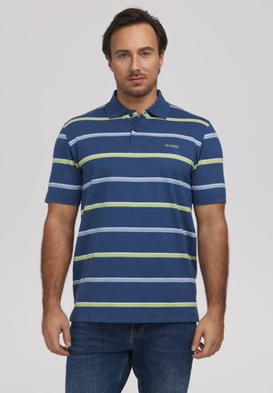 Mężczyzna w niebieskiej koszulce polo z zielonymi i jasnoniebieskimi poziomymi paskami, stojący na jednolitym jasnym tle.