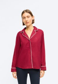 Bordová košeľa s dlhým rukávom a bielym lemovaním, s gombíkovým zapínaním a golierom. Vyrobená z mäkkej látky, má voľný strih a zošitú nášivku.