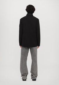 Zwarte jas met een hoge kraag, lange mouwen en een getailleerde taille, gecombineerd met losse grijze broek in een gestructureerde stof.