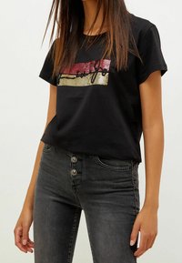 Kvinna som bär svart t-shirt med kort ärm med röda och guldsequinränder och mörkgrå högmidjade jeans med tre knappar.