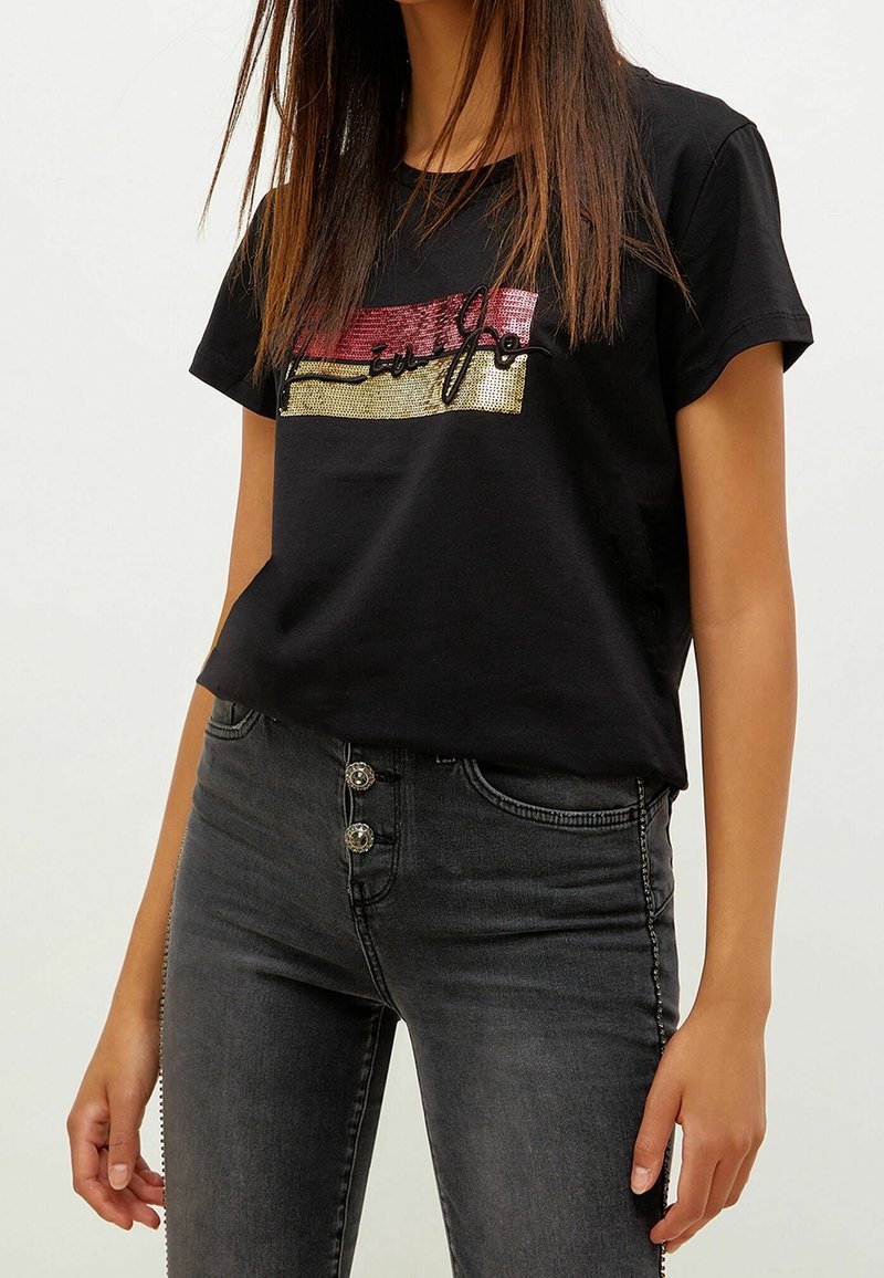 Kvinna som bär svart t-shirt med kort ärm med röda och guldsequinränder och mörkgrå högmidjade jeans med tre knappar.