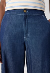 Blaue Denim-Hosen mit einer maßgeschneiderten Passform, ausgestattet mit einem Bauchknopf und kontrastierenden Nähten entlang der Nähte und Taschen. Glatte Textur.