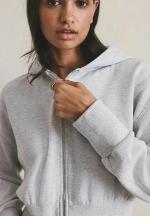 Femeie cu pistrui purtând un hanorac gri deschis cu fermoar, ținând fermoarul aproape de guler cu mâna dreaptă, fundal neutru.