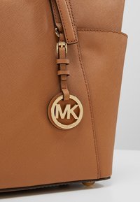 Sac cabas en cuir couleur tan avec poche latérale et pendentif logo en métal doré suspendu avec les initiales "MK".