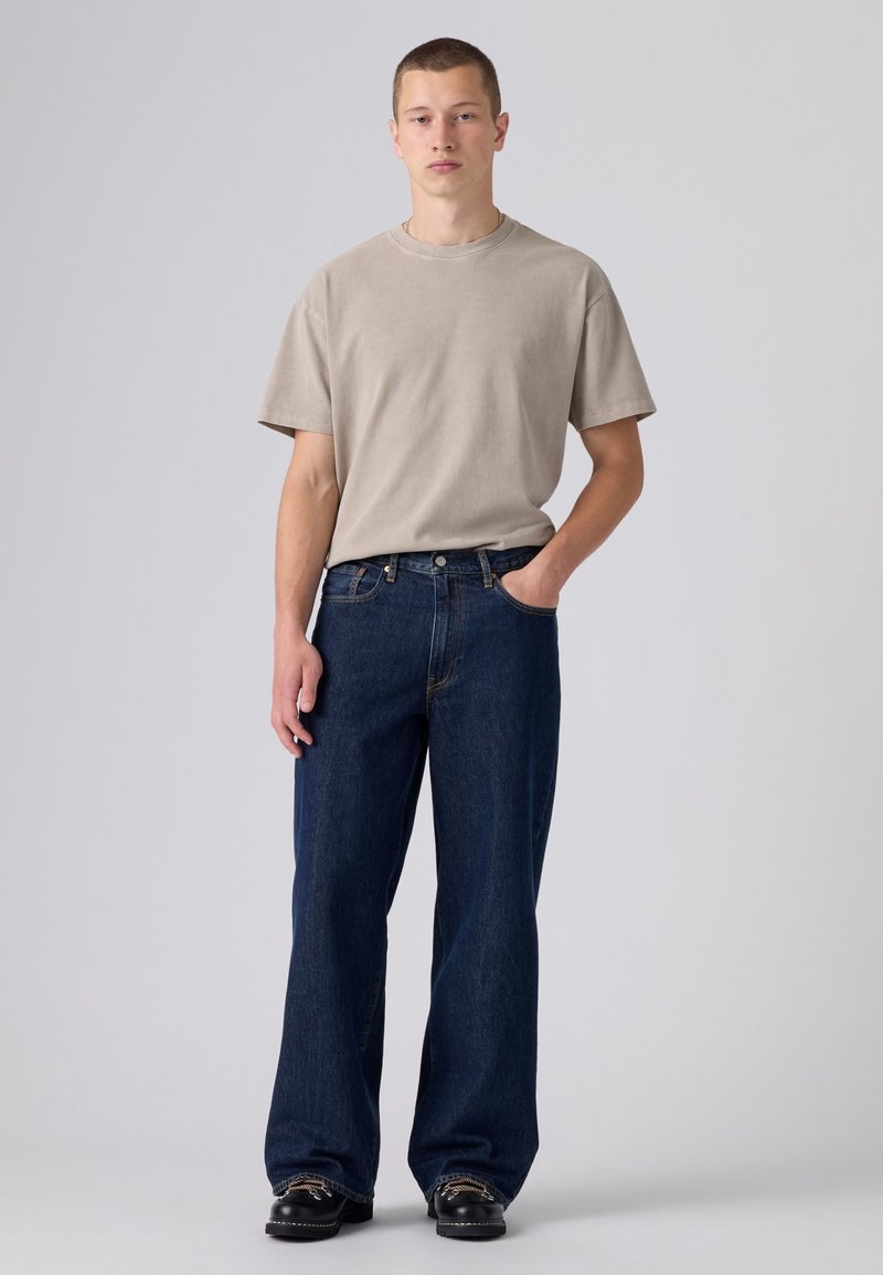 Jeune homme debout, une main dans la poche, portant un t-shirt beige, un jean large bleu foncé et des chaussures noires sur fond uni.