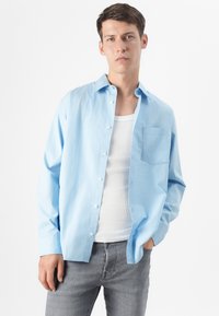 Ein junger Mann posiert selbstbewusst, trägt ein hellblaues Hemd über einem weißen Tanktop, graue Jeans und hat kurzes braunes Haar.