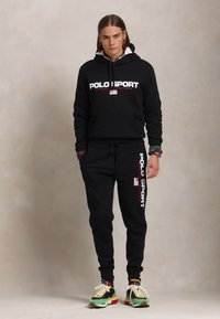 Melna kapuce un sporta bikses komplekts, kurā redzams "POLO SPORT" uzraksts baltā un sarkanā krāsā. Apģērbs ietver krāsainus sporta apavus ar dažādiem rakstiem.