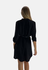 Robe noire à manches longues, taille cintrée et détail de poche arrière. Texture lisse, longueur genou, coupe décontractée.