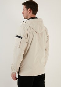 Beige waterdichte jas met een capuchon, ritszakken aan de zijkanten en een ritszak op de arm. Voorzien van elastische manchetten en een lichtgewicht ontwerp.