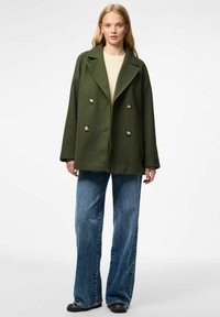 Olijfgroene dubbelzijdige wollen jas met gouden knopen, brede revers en relaxte pasvorm, gecombineerd met lichtblauwe flared jeans en zwarte schoenen.