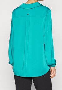 Turquoise blouse featuring a classic collar, button detail at the back, and elasticized cuffs. Smooth texture with a relaxed fit.