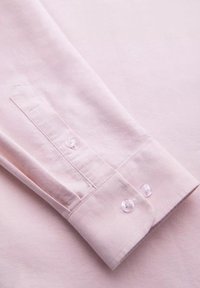 Poignet de chemise rose clair à manches longues avec deux boutons transparents et couture visible sur tissu lisse.
