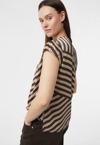 Frau mit langen braunen Haaren, die eine kurzärmelige beige und dunkelbraun gestreifte Bluse trägt, die Hände in den Taschen einer dunklen Hose, und über die Schulter schaut.