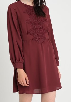 Femme portant une robe bordeaux à manches longues avec des détails en dentelle à l'avant et des poignets froncés aux manches.