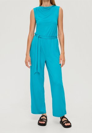 Turkusowy kombinezon z drapowanym dekoltem, szerokimi nogawkami, talią wysmukloną paskiem i miękką tkaniną. Noszony z czarnymi sandałami na platformie.