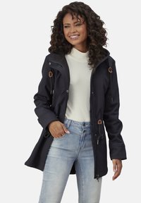 Navy softshell coat with a hood, drawstring details, and two front pockets, paired with a cream turtleneck and light blue jeans.