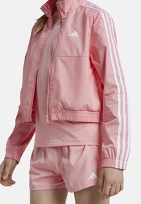 Chaqueta deportiva rosa con cremallera frontal y puños elásticos, que presenta acentos de tres rayas blancas en las mangas y pantalones cortos a juego.
