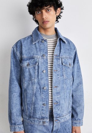 Giacca di jeans blu chiaro dal taglio classico, con due tasche sul petto, bottoni argentati e dettagli di cucitura in evidenza. Indossata sopra una camicia a righe.