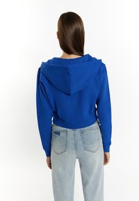 Felpa blu corta con cappuccio, maniche lunghe e orlo a coste. Abbinata a jeans in denim chiaro con tasche posteriori usurate.