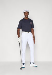 Camisola de manga curta azul-marinho com logótipo, calças brancas, cinto branco e sapatos brancos com detalhes azuis. A segurar um taco de golfe, a usar um boné branco.