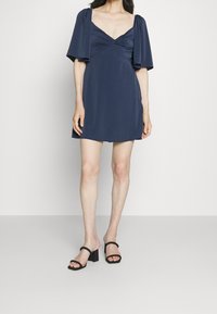 Granatowa krótka sukienka z dekoltem w serek, z bufkami na rękawach i dopasowanym talią. Noszona z czarnymi sandałami na obcasie w kształcie bloku. Gładka faktura.