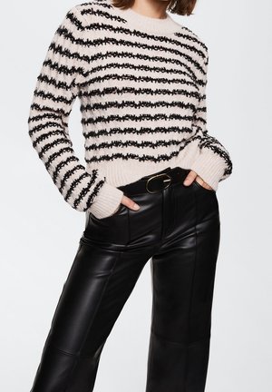 Femme portant un pull rayé crème et noir en matière pelucheuse avec des poignets côtelés, associé à un pantalon en cuir noir taille haute et une ceinture avec une boucle dorée.