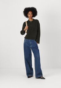 Dark green knitted cardigan with ribbed texture, front zipper, and two patch pockets. Paired with loose blue denim trousers and black flats.