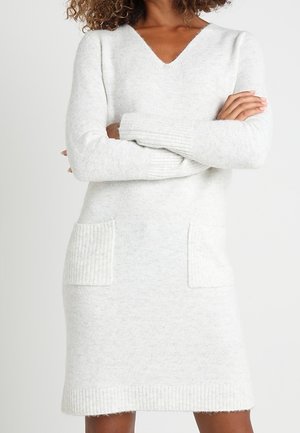 Vrouw draagt een wit gebreide jurk met lange mouwen, V-hals en twee zakken aan de voorkant, armen gekruist op de borst.