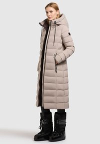 Lång, ljusbeige pufferjacka med ett kviltat mönster, fuskpälsfoder i huvan, svarta dragkedjor och sidofickor. Matchas med svarta isolerade stövlar.