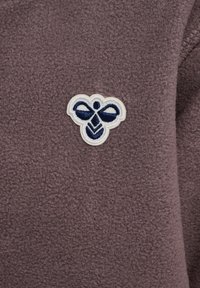 Bruin fleece stof met een zachte textuur, voorzien van een witte en marineblauwe geborduurde logo van een gestileerde bij. Geen zichtbare hardware of accenten.