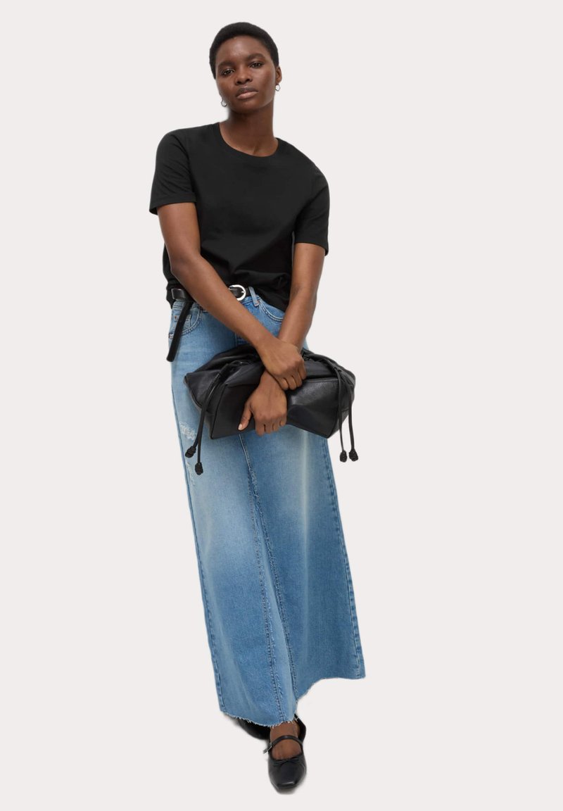 T-shirt nera, gonna di denim chiaro, tiene una borsa di pelle nera. La gonna presenta un taglio lungo e ampio; silhouette complessiva rilassata.