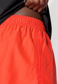 Κόκκινες σορτς με ελαστική μέση και πλαϊνή τσέπη, κατασκευασμένες από ελαφρύ ύφασμα. Ορατές λεπτομέρειες ραφής, συνδυασμένες με μαύρο πουκάμισο.