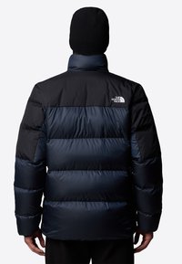 Mörkblå puffjacka med hög krage, med dunisolering, horisontellt kviltad design och en logotyp på övre ryggen.