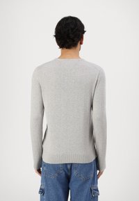 Personne aux cheveux courts et foncés portant un pull à manches longues gris clair et un pantalon cargo en denim bleu, vue de dos.