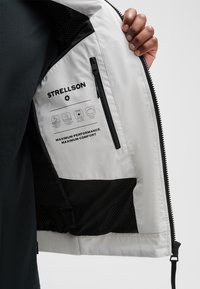 Weißes wasserdichtes Jäckchen mit schwarzen Akzenten, ausgestattet mit einer Reißverschlusstasche und Pflegehinweisen. Glatte Textur, leichtes Design und reisefreundlich.