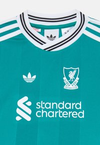 Turkizna nogometna majica s črno-belo prugasto ovratnikom, ki vsebuje grb Liverpool Football Club in logo Standard Chartered.