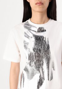 Hvid T-shirt med et stort metallic sølv abstrakt mønster på forsiden. Blødt bomuldsstof med rund halsudskæring og korte ærmer.