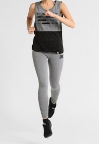 Woman running in gray sleeveless top, black mesh panel, gray leggings with logo, and black athletic shoes against plain background.