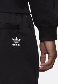 Černé sportovní tepláky s bočními kapsami na zip a bílým logem Adidas. Materiál má hladký povrch s mírným leskem.