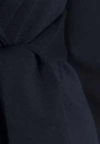 Černá, elastická tkanina s hladkou texturou, s uzlovým detailem na místech, kde se materiál překrývá. Detailní záběr na design.
