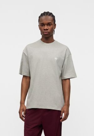 Ung mand med cornrows iført en løs lysegrå t-shirt og mørkerøde bukser, står mod en ensfarvet hvid baggrund.