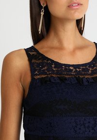 Haut débardeur en dentelle bleu marine avec un design en dentelle superposée, des accents à volants, un col rond et des panneaux texturés noirs contrastants.