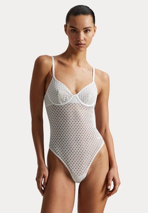 Frau mit dunklem Haar, die einen weißen Body mit Spitzenmuster und dünnen Trägern trägt, steht vor einem einfarbigen Hintergrund.