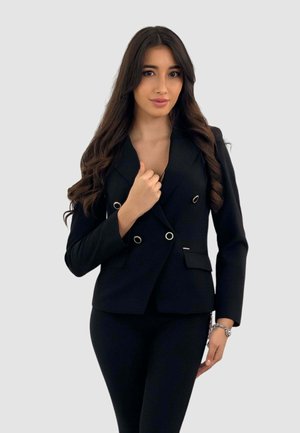 Blazer nero doppiopetto con dettagli in metallo, design strutturato, rever a scotto e tasche laterali, indossato sopra pantaloni neri aderenti.